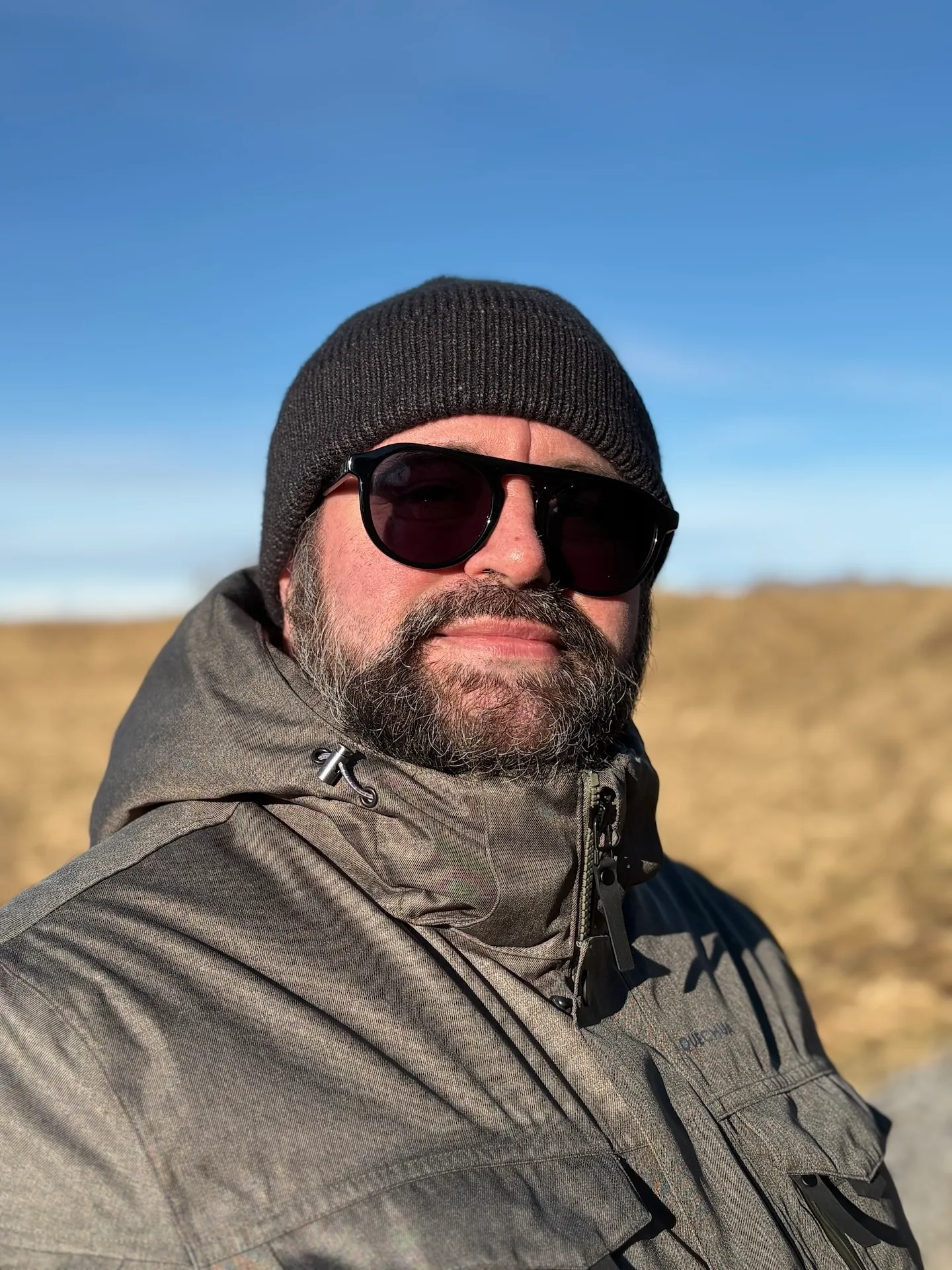 Raphaël Lemaitre in Iceland, wearing a beanie, sunglasses and a parka, in front of dry grass and a blue sky